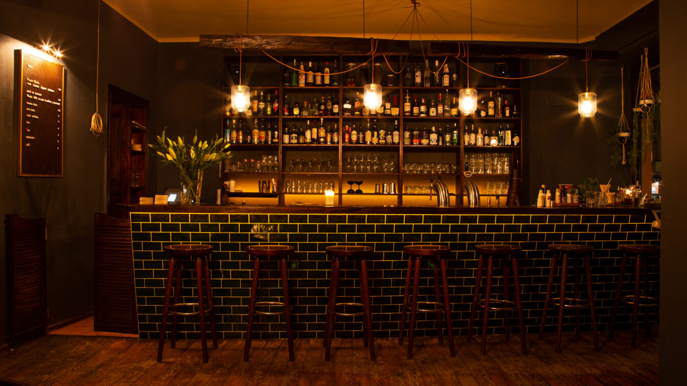 Foto von einem Bartresen mit grünen Fließen und Barhockern aus Holz davor. Drei Glühbirnen in der Bar in Berlin sorgen für eine gedimmte Lichtstimmung.